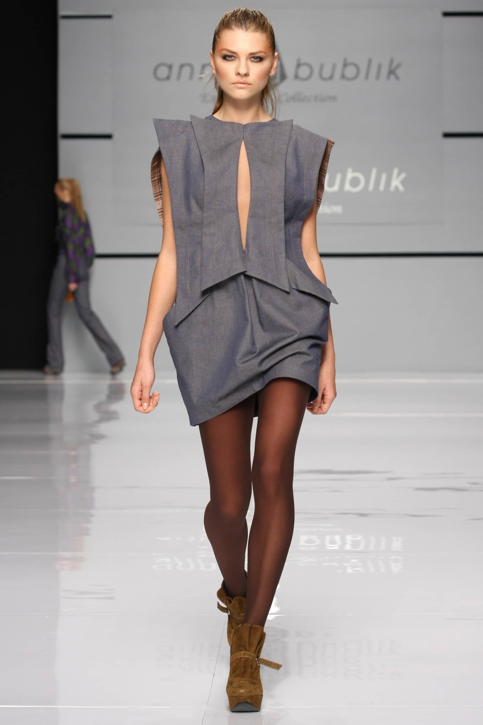 A model walks the runway wearing a gray sleeveless mini dress with a deep keyhole cutout and layered ruffle details, paired with sheer black tights and brown suede ankle boots. The background shows the 'Anna Bublik' brand name on the runway wall.
