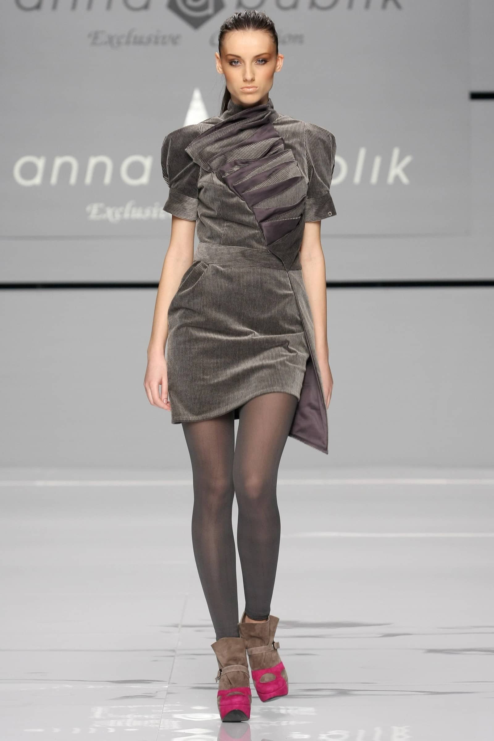 A runway model walks the catwalk wearing an Anna Blik Exclusive fashion dress. The dress is a short, structured, asymmetrical mini dress in a dark brown corduroy fabric with a high draped collar and layered ruffle details on the chest.