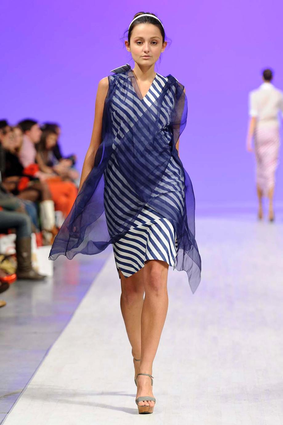 A model walks down a fashion runway wearing a sleeveless, V-neck dress with navy and white diagonal stripes, draped with a matching sheer navy wrap, against a purple runway backdrop.